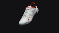 A sleek white sports shoe with red accents and logo details, featuring a textured surface and clean design lines. The background is dark, emphasizing the shoe.