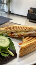 A grilled sandwich with melted cheese and sliced ingredients is placed on a white plate, accompanied by fresh green leaves and cucumber slices. The background features a modern interior with a sofa and plant, adding a cozy and inviting ambiance.