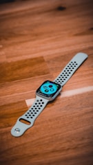 A smartwatch with a perforated white strap lies on a wooden surface. The screen displays a colorful interface with various icons and complications.