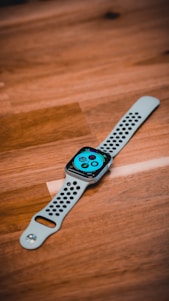 A smartwatch with a perforated white strap lies on a wooden surface. The screen displays a colorful interface with various icons and complications.