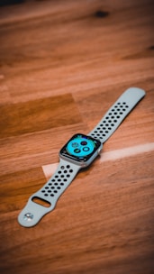 A smartwatch with a perforated white strap lies on a wooden surface. The screen displays a colorful interface with various icons and complications.