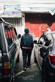 A person in a dark jacket and hat walks between parked cars on a street with a textured, red metal door in the background. Overhead, several signs are visible, with one saying 'Sahabat Keluarga Anda.'