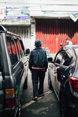 A person in a dark jacket and hat walks between parked cars on a street with a textured, red metal door in the background. Overhead, several signs are visible, with one saying 'Sahabat Keluarga Anda.'