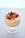 A dessert in a glass featuring layers of creamy white yogurt or mousse, topped with bright yellow mango pieces, fresh red raspberries, and grated chocolate. The glass is set on a light, neutral surface with soft lighting that highlights the dessert's textures and colors.