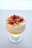 A dessert in a glass featuring layers of creamy white yogurt or mousse, topped with bright yellow mango pieces, fresh red raspberries, and grated chocolate. The glass is set on a light, neutral surface with soft lighting that highlights the dessert's textures and colors.