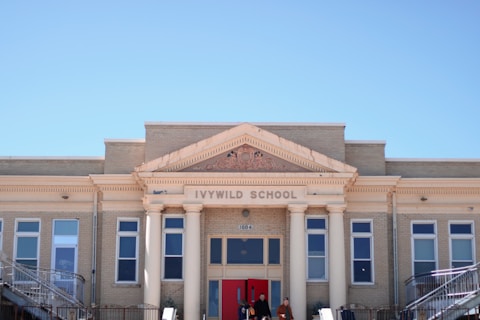 An old brick school building with classic architectural features, including columns and decorative details around the entrance. The facade displays the name 'Ivywild School' above the entrance, with several large windows aligned symmetrically on either side. Two people are visible at the entrance.
