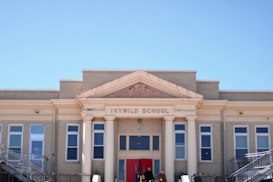 An old brick school building with classic architectural features, including columns and decorative details around the entrance. The facade displays the name 'Ivywild School' above the entrance, with several large windows aligned symmetrically on either side. Two people are visible at the entrance.