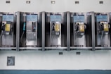 A row of vintage public payphones is mounted on a tiled wall. The payphones are metallic with varying colors, including yellow and black. They are spaced evenly and appear to be part of an older communication infrastructure.