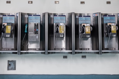 A row of vintage public payphones is mounted on a tiled wall. The payphones are metallic with varying colors, including yellow and black. They are spaced evenly and appear to be part of an older communication infrastructure.
