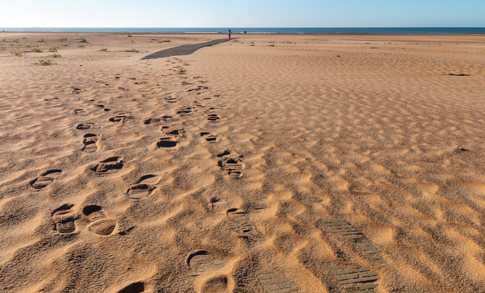 Playa de arena amplia con huellas y una pasarela de madera al fondo, en la costa de Huelva