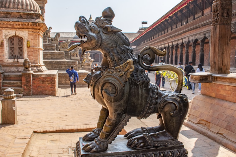 A detailed bronze statue of a mythical creature sits prominently in an ancient courtyard, surrounded by historic, intricately carved stone structures. People are walking around, with one person wearing vibrant clothing standing near the sculpture. The area is bathed in sunlight, creating a warm and historic atmosphere.