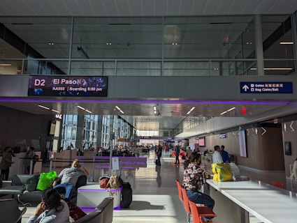 A busy airport terminal with several people seated and walking around. Overhead digital signage shows a flight to El Paso at gate D2. The terminal has high ceilings and natural light coming through the large windows. Some people are sitting on chairs or couches, with bags around them, while others are moving through the space.