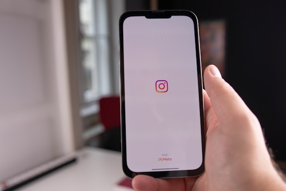 A person holding a smartphone, displaying the Instagram logo on its screen. The surrounding area appears to be indoors, featuring natural light coming through a window and blurry elements in the background.
