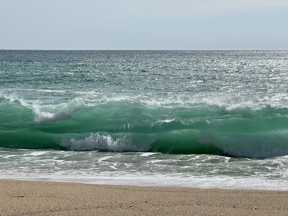 Large wave crashes onto sandy shore with white foam and green water in background