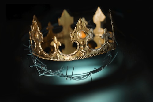 An ornate gold crown with intricate designs and jewel-like accents sits atop a circular piece of barbed wire. The image is set against a dark background, highlighting the contrasting elements of regality and harshness.