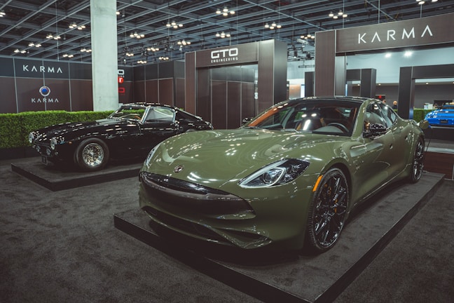 Two luxury cars are displayed in an indoor showroom setting with modern lighting and company branding. The foreground features a green sports car with sleek, aerodynamic design and polished black wheels. In the background, a black classic car with distinctive chrome details is positioned on display.