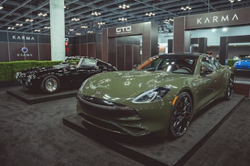 Two luxury cars are displayed in an indoor showroom setting with modern lighting and company branding. The foreground features a green sports car with sleek, aerodynamic design and polished black wheels. In the background, a black classic car with distinctive chrome details is positioned on display.