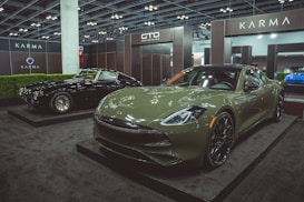 Two luxury cars are displayed in an indoor showroom setting with modern lighting and company branding. The foreground features a green sports car with sleek, aerodynamic design and polished black wheels. In the background, a black classic car with distinctive chrome details is positioned on display.