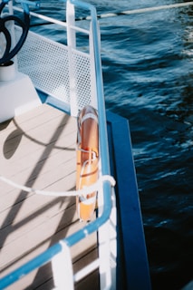 The image captures a section of a boat's deck bordered by a white metal railing with blue accents. The deck appears wooden, and attached to the railing is an orange lifebuoy. The boat is on a body of water, visible on the right side of the image, which is deep blue and gently rippling.