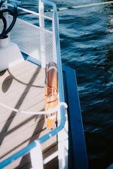 The image captures a section of a boat's deck bordered by a white metal railing with blue accents. The deck appears wooden, and attached to the railing is an orange lifebuoy. The boat is on a body of water, visible on the right side of the image, which is deep blue and gently rippling.