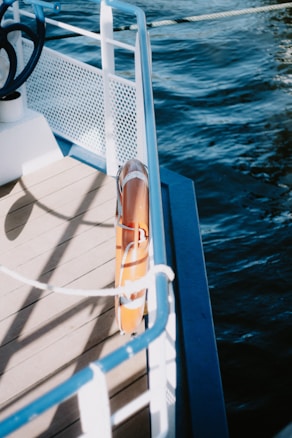 The image captures a section of a boat's deck bordered by a white metal railing with blue accents. The deck appears wooden, and attached to the railing is an orange lifebuoy. The boat is on a body of water, visible on the right side of the image, which is deep blue and gently rippling.