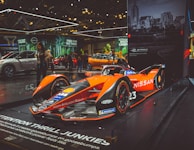 A sleek, orange and black Formula E race car displayed prominently on a raised platform at an auto show. The car is branded with Nissan and various other sponsor logos. Surrounding the car are other vehicles and a few people examining the exhibits. Large banners with advertising slogans hang in the background, along with an urban skyline image.