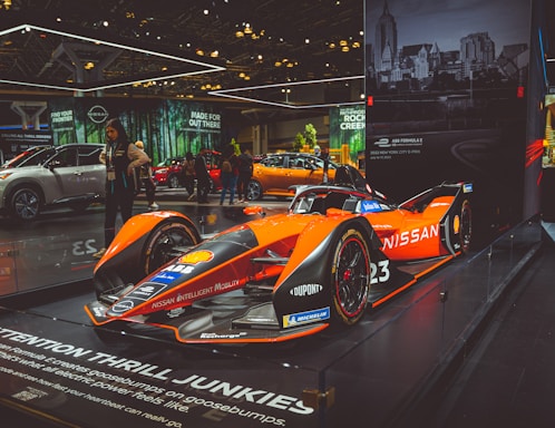 A sleek, orange and black Formula E race car displayed prominently on a raised platform at an auto show. The car is branded with Nissan and various other sponsor logos. Surrounding the car are other vehicles and a few people examining the exhibits. Large banners with advertising slogans hang in the background, along with an urban skyline image.