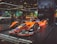 A sleek, orange and black Formula E race car displayed prominently on a raised platform at an auto show. The car is branded with Nissan and various other sponsor logos. Surrounding the car are other vehicles and a few people examining the exhibits. Large banners with advertising slogans hang in the background, along with an urban skyline image.