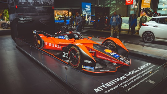 A sleek, modern race car with a bright orange and black color scheme displayed prominently on a show floor. Various brand logos such as Nissan, Shell, and Michelin are visible on the car. The backdrop includes a few other vehicles and people examining the displays. The area is well-lit with concentrated spotlights highlighting the car.
