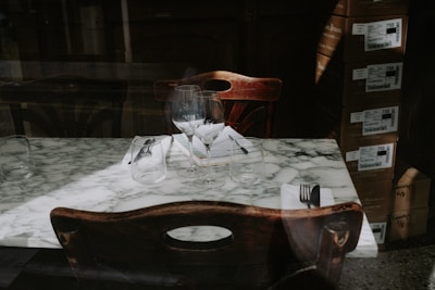 An elegant marble table set for two with wine glasses and utensils rests against a dark wooden backdrop. Boxes are stacked in the corner, adding an industrial element to an otherwise refined ambiance.