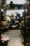 A cozy, indoor flower shop filled with a variety of potted plants and flowers. The space is densely arranged with large pots on the floor and tables, hosting lush green foliage and colorful blossoms. Soft, natural light streams in, creating a serene atmosphere. The walls are adorned with hanging plants, and the floor tiles lend a rustic charm.