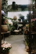 A cozy, indoor flower shop filled with a variety of potted plants and flowers. The space is densely arranged with large pots on the floor and tables, hosting lush green foliage and colorful blossoms. Soft, natural light streams in, creating a serene atmosphere. The walls are adorned with hanging plants, and the floor tiles lend a rustic charm.