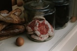 A variety of baked goods are displayed in glass jars on a counter. Large, rustic baguettes rest alongside jars filled with colorful meringue cookies, featuring red and white swirls. A couple of brown eggs sit nearby, adding a rustic touch to the scene.
