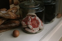 A variety of baked goods are displayed in glass jars on a counter. Large, rustic baguettes rest alongside jars filled with colorful meringue cookies, featuring red and white swirls. A couple of brown eggs sit nearby, adding a rustic touch to the scene.