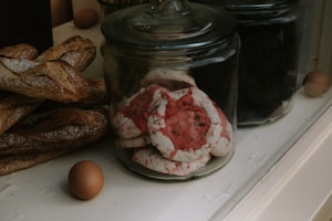 A variety of baked goods are displayed in glass jars on a counter. Large, rustic baguettes rest alongside jars filled with colorful meringue cookies, featuring red and white swirls. A couple of brown eggs sit nearby, adding a rustic touch to the scene.
