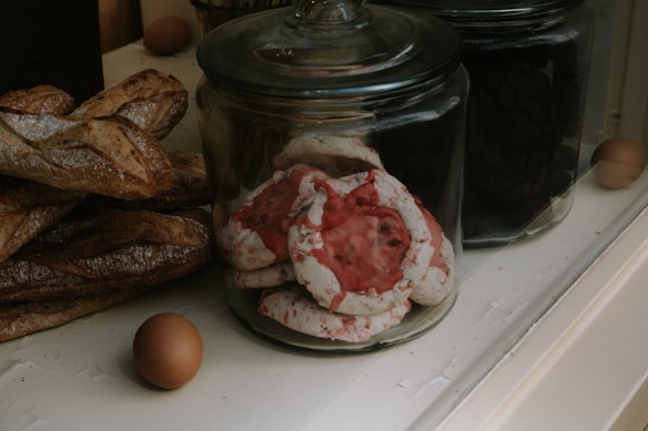 A variety of baked goods are displayed in glass jars on a counter. Large, rustic baguettes rest alongside jars filled with colorful meringue cookies, featuring red and white swirls. A couple of brown eggs sit nearby, adding a rustic touch to the scene.