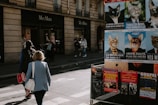 Pedestrians walk near a MaxMara store on a street lined with advertisement posters featuring cats in humorous political attire. The storefront has mannequins on display, and there are various posters promoting concerts and events on a nearby billboard. The scene has a candid, urban vibe with people casually going about their day.