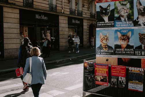 Pedestrians walk near a MaxMara store on a street lined with advertisement posters featuring cats in humorous political attire. The storefront has mannequins on display, and there are various posters promoting concerts and events on a nearby billboard. The scene has a candid, urban vibe with people casually going about their day.