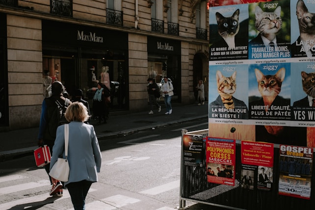 Pedestrians walk near a MaxMara store on a street lined with advertisement posters featuring cats in humorous political attire. The storefront has mannequins on display, and there are various posters promoting concerts and events on a nearby billboard. The scene has a candid, urban vibe with people casually going about their day.