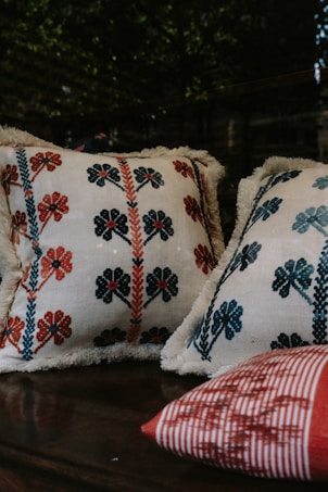 Four pillows featuring embroidered floral patterns are placed together. Two of the pillows have intricate designs with blue and red flowers, while one has a red and white striped pattern. The background is dark, making the pillows stand out.