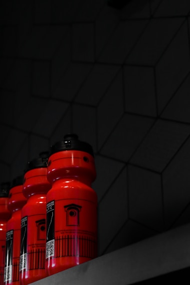 A set of bright red water bottles with black caps are neatly arranged on a shelf against a dark, geometric-patterned wall. The bottles feature a black graphic design, including text and a QR code.
