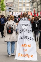 People are participating in a protest march on a city street lined with buildings. One individual is wearing a long cloth with messages written in German, accompanied by another person with a backpack featuring an orange cross. The crowd is holding banners and red flags, suggesting a political or social movement.
