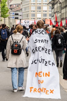 People are participating in a protest march on a city street lined with buildings. One individual is wearing a long cloth with messages written in German, accompanied by another person with a backpack featuring an orange cross. The crowd is holding banners and red flags, suggesting a political or social movement.
