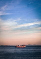 A large container ship sails across a calm sea, with red and orange shipping containers stacked on its deck. The horizon reveals a distant mountain range under a soft pastel sky transitioning from blue at the top to warm hues near the horizon line.