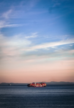 A large container ship sails across a calm sea, with red and orange shipping containers stacked on its deck. The horizon reveals a distant mountain range under a soft pastel sky transitioning from blue at the top to warm hues near the horizon line.