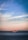 A large container ship sails across a calm sea, with red and orange shipping containers stacked on its deck. The horizon reveals a distant mountain range under a soft pastel sky transitioning from blue at the top to warm hues near the horizon line.