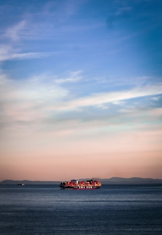 A large container ship sails across a calm sea, with red and orange shipping containers stacked on its deck. The horizon reveals a distant mountain range under a soft pastel sky transitioning from blue at the top to warm hues near the horizon line.