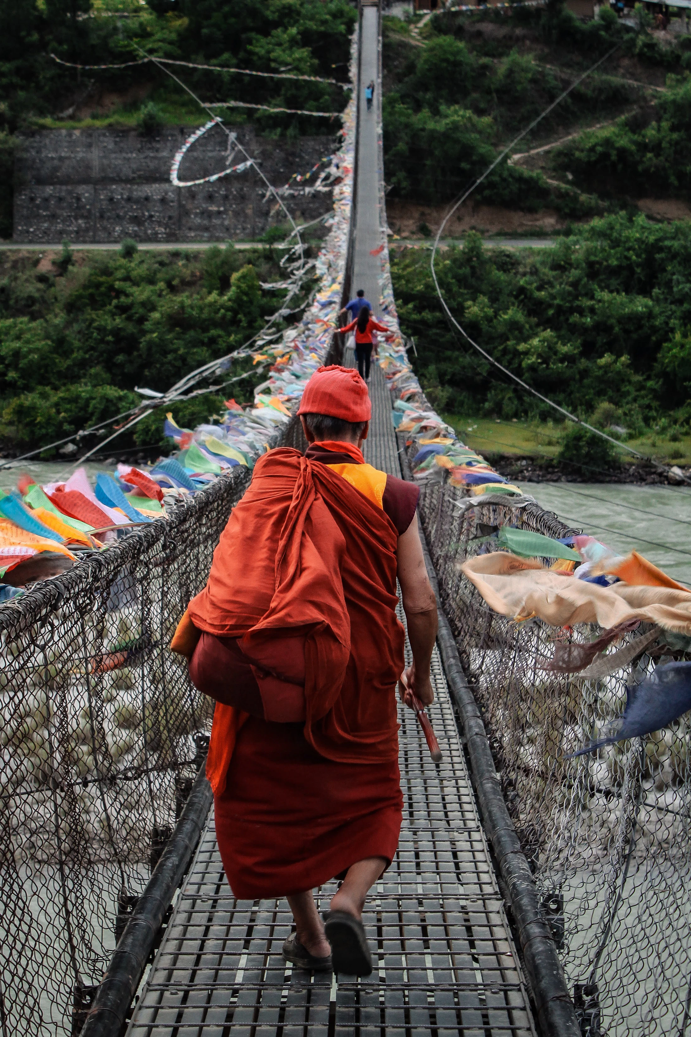 A monk in saffron crossing a prayer-flag-strung suspension bridge