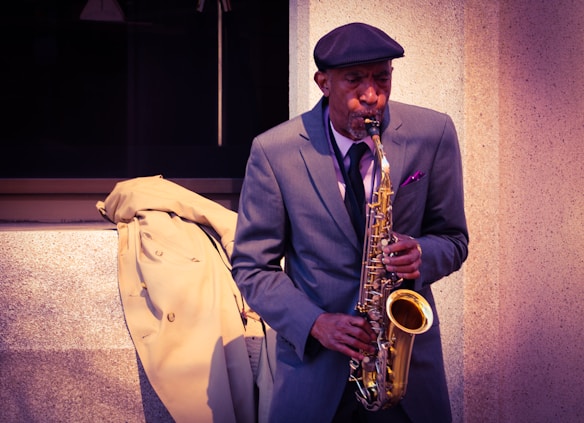 A man in a suit is playing a saxophone on the street, with a beige coat draped over the ledge beside him. He stands against a textured concrete wall, wearing a flat cap and a pocket square visible in his suit jacket.