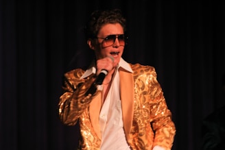 A person is performing on stage, wearing a shiny gold jacket with intricate patterns, a white shirt, and large sunglasses. The individual is holding a microphone and is captured in mid-performance, conveying energy and enthusiasm.